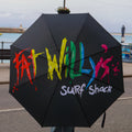 Black umbrella with colorful Fat Willy's logo held by a person on a coastal road.